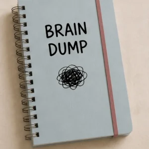Libreta de espiral con tapas de color azul grisáceo y banda elástica rosa. En la portada se lee "BRAIN DUMP" sobre el dibujo de un garabato negro.